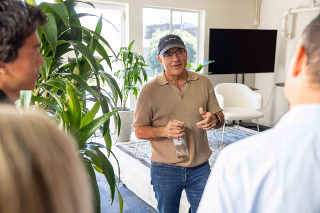 A man in a brown polo and black cap talks to three people in a bright room with green plants, a white chair, and a large TV screen.
